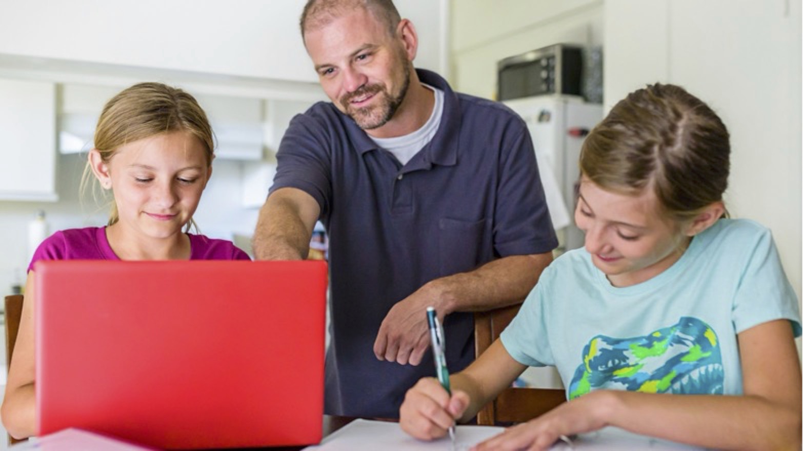 An adult in a navy polo shirt helps two young students with schoolwork at a table. One student is looking at a red laptop while the other writes with a pen. The students are wearing casual clothes - one in pink/magenta and one in light blue with a dinosaur design. The scene appears to be in a kitchen or study area with white cabinets visible in the background.